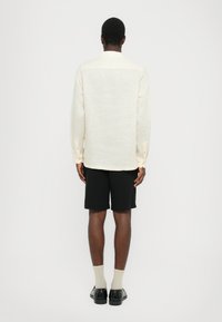 Lysbeige langermet skjorte med teksturert overflate, knappelukking ved ermene og buet søm, kombinert med svarte shorts og svarte sko.
