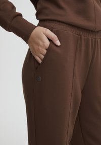 Bruna bomullssweatpants med texturerad yta, elastisk midja och sidofickor. Har en metall-logotyp som detalj nära fickan.