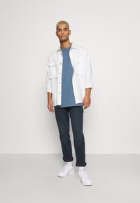 Camisa blanca de botones, camiseta azul, jeans oscuros y zapatillas blancas. El modelo está de pie en un fondo neutro, mostrando un atuendo casual.