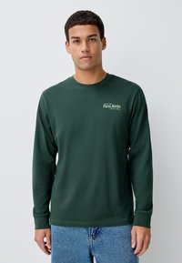 Langærmet t-shirt lavet af mørkegrønt bomuld med rund halsudskæring, lige snit og et lille hvidt tryk med teksten "Paris.Berlin." på brystet.