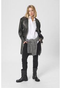 Nő áll, kezét a zsebében tartja, fekete bőrdzsekit visel fehér inget, derekán szürke pulóvert kötve, fekete nadrágot és fekete csizmát.