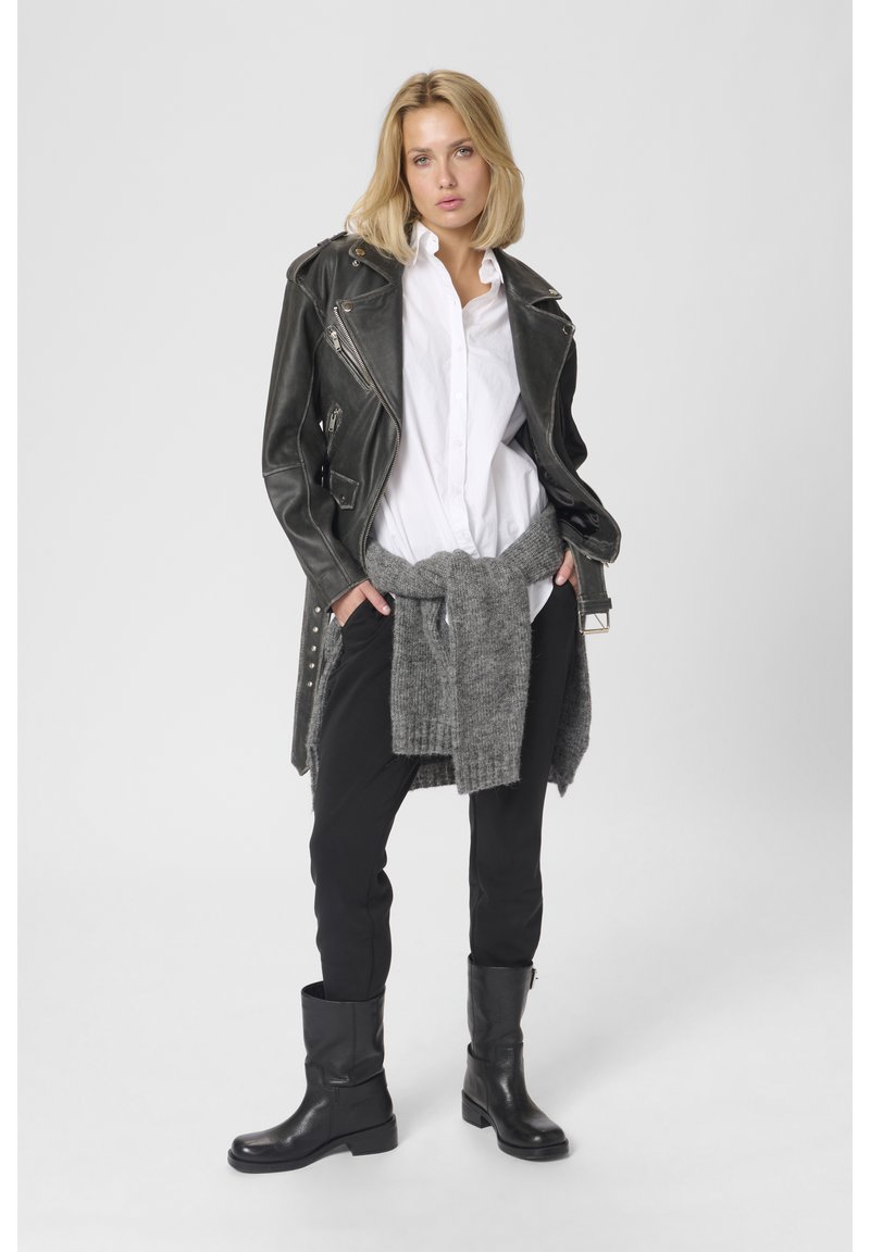 Nő áll, kezét a zsebében tartja, fekete bőrdzsekit visel fehér inget, derekán szürke pulóvert kötve, fekete nadrágot és fekete csizmát.