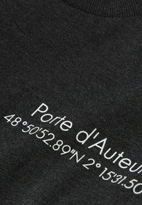 Melns kokvilnas audums ar baltu tekstu "Porte d'Auteuil" un ģeogrāfiskajām koordinātēm, tuvināts gludas struktūras skats.