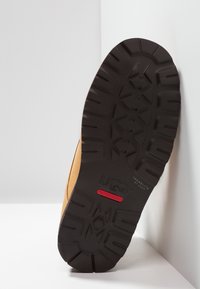 Suela de bota impermeable UGG marrón inclinada contra una esquina blanca, mostrando el patrón de tracción negro y la etiqueta impermeable roja.