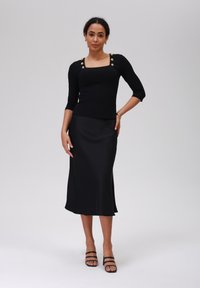 Zwarte geribbelde longsleeve top met vierkante halslijn en gouden knopenaccenten, gecombineerd met een slanke zwarte satijnen midi-jurk en zwarte enkellaarsjes met bandjes.