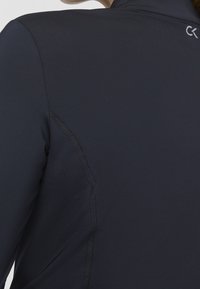 Detailní záběr osoby v tmavě modrém sportovním topu se viditelným švem a malým bílým logem CK blízko límce.