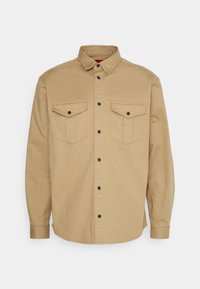 Camisa de manga larga beige con botones, dos bolsillos frontales con solapa y botones, un collar, y el logo "HUGO" bordado en el bolsillo derecho.