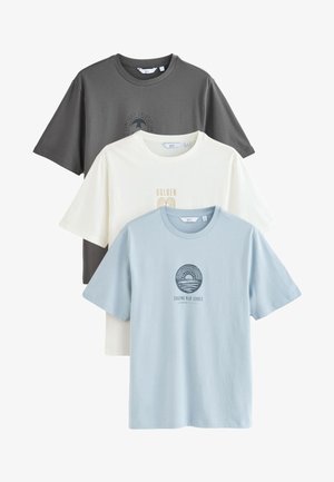 Trois t-shirts à col rond pliés en gris, crème et bleu clair avec des imprimés graphiques centrés et manches courtes, superposés sur un fond blanc.