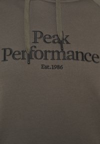 Sweat à capuche vert olive avec les inscriptions "Peak Performance" et "Est. 1986" brodées en noir. Dispose d'une capuche avec cordon de serrage et d'un tissu texturé.
