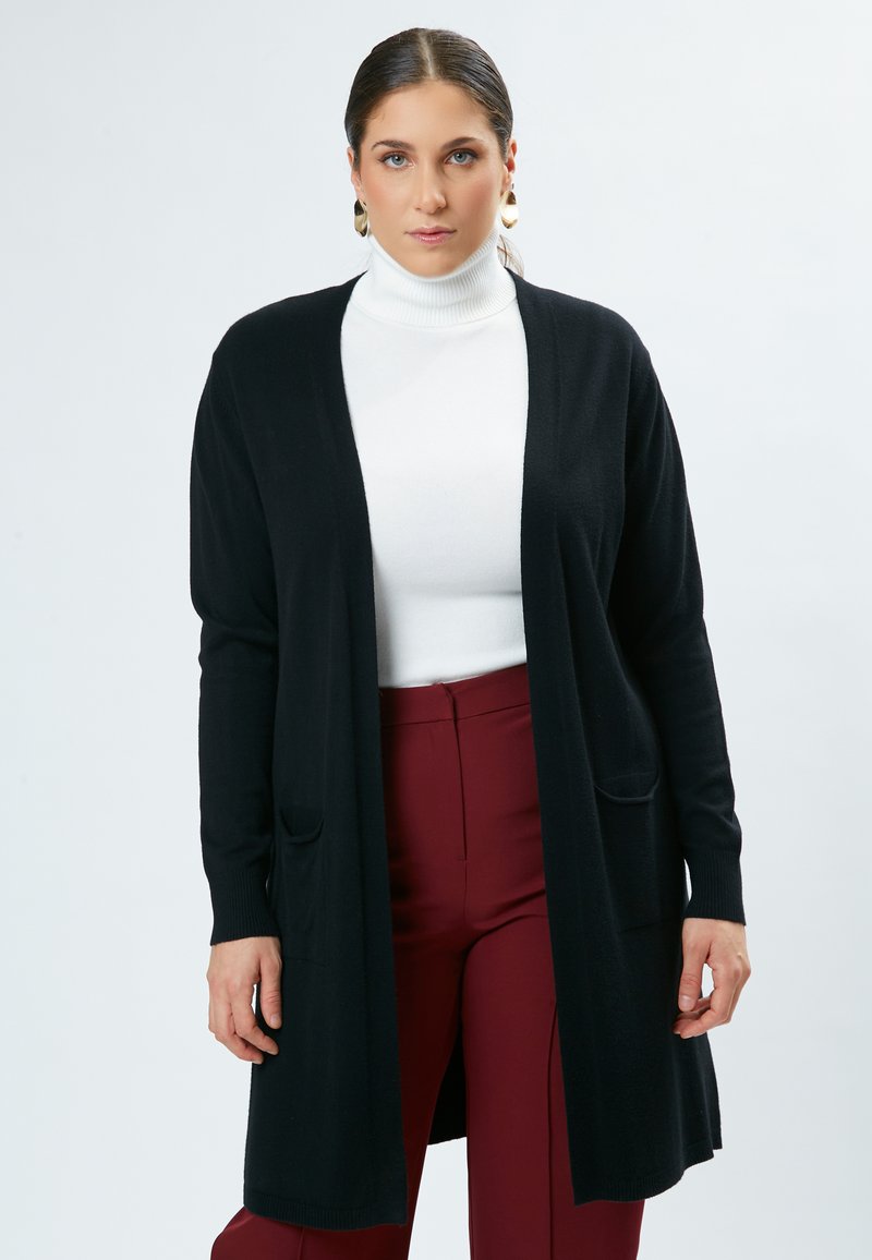 Zwarte lange cardigan van zacht materiaal, met een losse pasvorm, open voorkant, twee zakken en geribbelde manchetten. Gecombineerd met een witte coltrui en bordeauxrode broek.