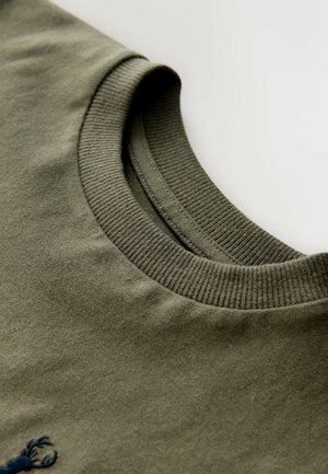 Olivezelené bavlnené tričko s rebrovaným okrúhlym výstrihom a hladkou, jemnou textúrou. Malý vyšívaný detail blízko spodnej časti.