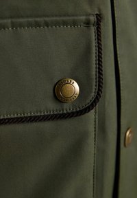 Olivgrüne Stofftasche mit schwarzem geflochtenem Besatz und einem runden Messingknopf mit der Aufschrift "Joules Est. 1989" an einem Kleidungsstück.