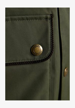 Olive green fabric pocket with black braided trim and a round brass button labeled "Joules Est. 1989" on a garment.
