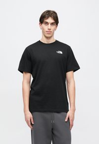 Μαύρο βαμβακερό μπλουζάκι με μεγάλο λευκό λογότυπο στο αριστερό μέρος του στήθους, κοντά μανίκια και κλασική στρογγυλή λαιμόκοψη. Φορεμένο με γκρι παντελόνι.