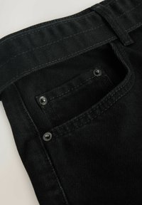 Schwarzer Denimstoff mit einer detaillierten Nahaufnahme der Gesäßtasche und des Bunds, mit sichtbaren Nähten und Metallnieten.