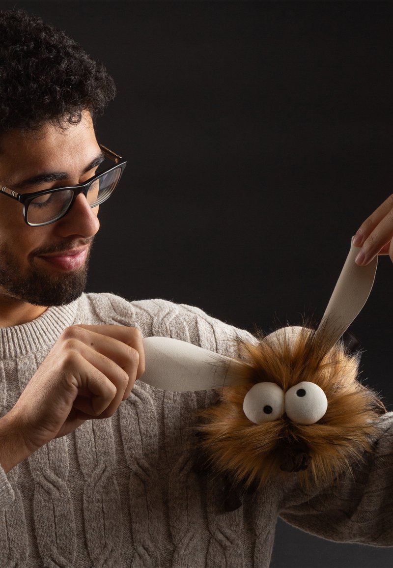 Brown furry puppet with large eyes and black features, held by a person wearing a cable-knit sweater, white ears attached.
