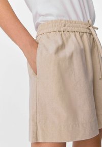 Pantaloni corti in lino beige con vita elastica e cordoncino, dotati di tasche laterali e dalla vestibilità comoda, che mostrano una texture liscia.