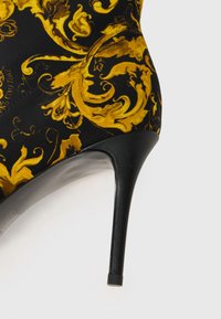 Zwarte hoge hak met een glanzende afwerking, voorzien van een geel en gouden sierlijk patroon op stof, puntige neus en slanke stiletto hak.