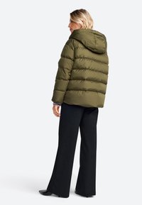 Donkergroene pufferjack met horizontale stiksel, aangehechte capuchon en een ontspannen pasvorm, gecombineerd met zwarte wijde broek en zwarte schoenen.