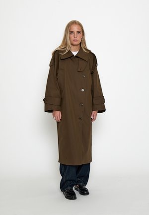 Junge Frau mit langen blonden Haaren trägt einen braunen übergroßen Trenchcoat über dunklen Jeans und schwarzen Schuhen, stehend vor weißem Hintergrund.