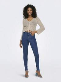 Γυναίκα στέκεται με το χέρι στην τσέπη φορώντας μπεζ πλεκτό κοντό πουλόβερ, μπλε skinny jeans και μπλε ψηλοτάκουνα πέδιλα μπροστά σε λευκό φόντο.