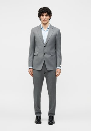 Άνδρας που στέκεται με το πρόσωπο προς τα εμπρός, φορώντας γαλάζιο πουκάμισο και γκρι κοστούμι με δύο κουμπιά, με μαύρα παπούτσια σε λευκό φόντο.