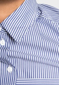 Modrá a bílá košile s vertikálními pruhy, klasickým límečkem, zaoblenými bílými knoflíky a hladkou, jemnou texturou látky.