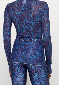 Pitkähihainen aktiivivaatetoppi purppuran ja sinisen eläinkuosin kanssa. Ominaisuuksiin kuuluu venyvä kangas, jossa on sileä rakenne ja heijastava logo-yksityiskohta.