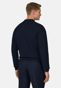Homme de dos portant un pull côtelé bleu marine foncé et un pantalon ajusté assorti, sur un fond clair uni.