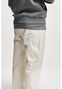 Persoon in beige cargobroek en een donkergrijze sweatshirt, weergegeven van de taille naar beneden tegen een effen achtergrond.