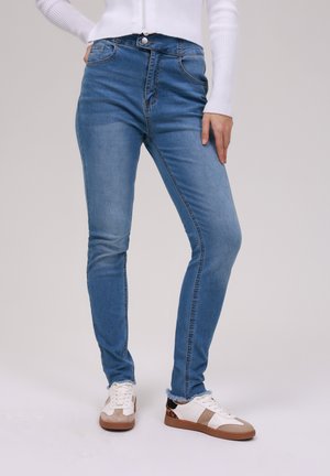 Blue skinny jeans made of denim with a mid-rise waist, five pockets, and a subtle faded finish; paired with white and beige sneakers.