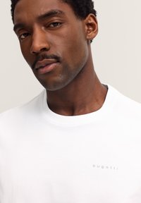 Nahaufnahme eines Mannes mit kurzen lockigen Haaren und einem Schnurrbart, der ein weißes Bugatti-Logo-T-Shirt trägt und leicht zur Seite schaut, vor einem beigen Hintergrund.