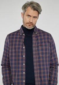 Koszula w kratę w granatowym, czerwonym i zielonym kolorze, z kołnierzykiem z guzikami, kieszenią na piersi oraz kontrastowymi białymi guzikami, noszona na ciemnym golfie.