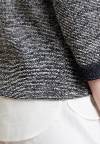 Ciemnoniebieski sweter z białym wzorem o teksturowanej fakturze, z okrągłym dekoltem i podwiniętymi mankietami, połączony z białym elementem odzieży z zaokrąglonymi akcentami na dole.