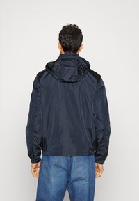 Giacca a vento blu navy in materiale leggero con una texture liscia; polsini e orlo elasticizzati; presenta dettagli sulle spalle e ai lati.