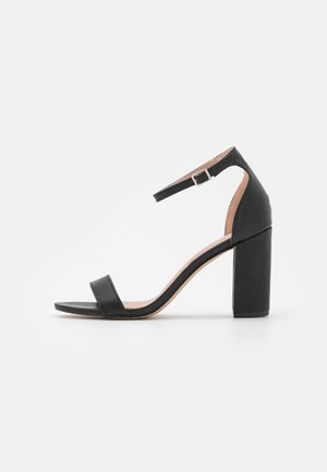 Czarny skórzany sandał na wysokim obcasie z szerokim, blokowym obcasem, regulowanym paskiem na kostkę, odkrytymi palcami i gładkim wykończeniem.