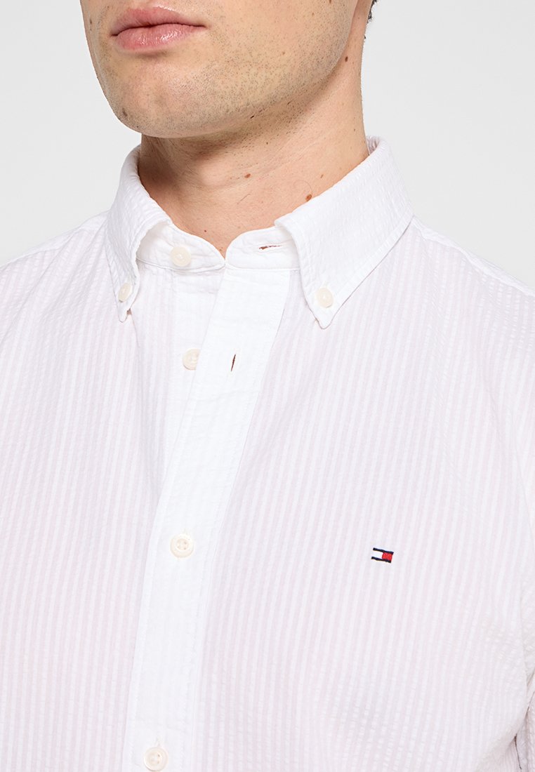 Gros plan d'une personne portant une chemise blanche à boutons avec un petit logo rouge, blanc et bleu sur la poitrine.