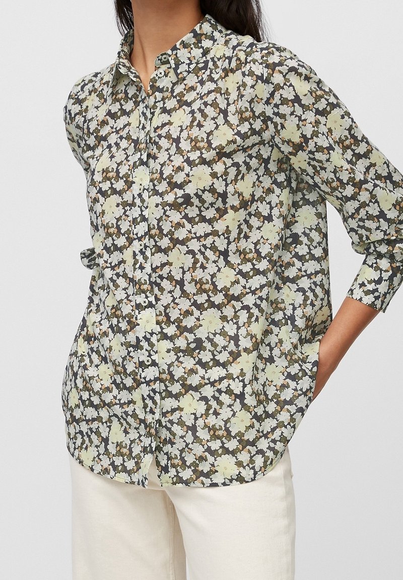 Femme portant une chemise boutonnée à manches longues avec un motif floral jaune et blanc associée à un pantalon blanc, posant avec une main dans la poche.