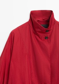 Rote leichte Jacke mit hohem Kragen, ausgestattet mit einer strukturierten Oberfläche und zwei schwarzen Knöpfen in der Nähe des Halses. Minimalistisches Design, keine auffälligen Muster.