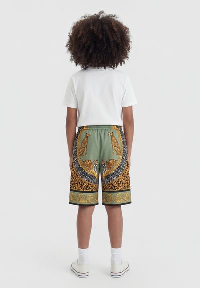 Enfant aux cheveux bouclés, debout de dos, portant un t-shirt blanc et un short à motifs avec des léopards et des tigres sur un fond vert et doré.