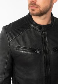 Giacca in pelle nera con colletto rialzato, dettagli cavi sulle spalle e tasche con zip frontali dotate di accessori neri.
