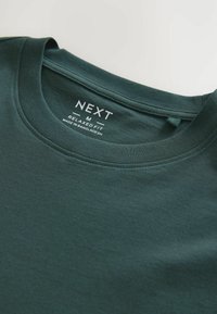 Tricou din bumbac verde închis, cu guler rotund, croială lejeră și etichetă imprimată care afișează informații despre marcă și îngrijire, plasată în interiorul gulerului.