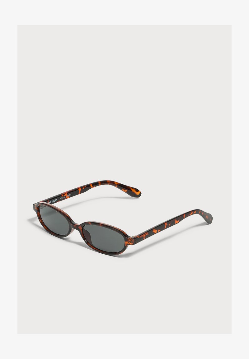 Petites lunettes de soleil ovales en écaille avec des verres foncés reposant sur une surface gris clair.