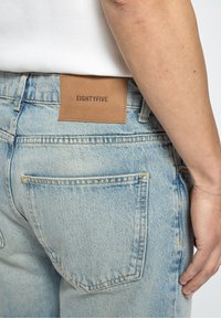 Jean en denim bleu clair avec une texture usée, comportant une étiquette en cuir marron marquée "EIGHTYFIVE" sur la ceinture et deux poches arrière.