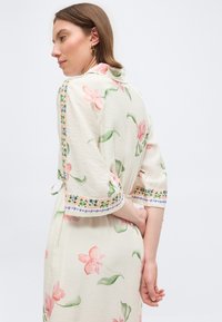 Γυναίκα με κοντά καστανά μαλλιά, ντυμένη με κρεμ φόρεμα με ροζ ανθικό μοτίβο και διακοσμητική πράσινη κεντημένη τρέσα, όρθια με τα χέρια πίσω από την πλάτη.