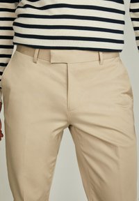 Pantaloni beige con una texture liscia e design a frontale piatto. Presentano un passante per cintura e un sottile dettaglio di cucitura frontale.