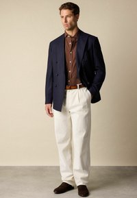 Marineblauwe pinstriped blazer over een bruine button-up shirt, gecombineerd met crèmekleurige wijde broek en donkerbruine loafers. Een eenvoudige riem accentueert de taille.