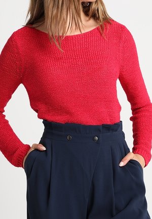 Vrouw draagt een rood, gestructureerd, lange-mouwen shirt ingestopt in een hooggetailleerde marineblauwe broek met handen in de zakken.