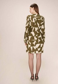 Robe à motifs olive et crème avec des manches longues, taille élastique et un détail plissé à l'arrière, associée à des escarpins noirs à lanières.