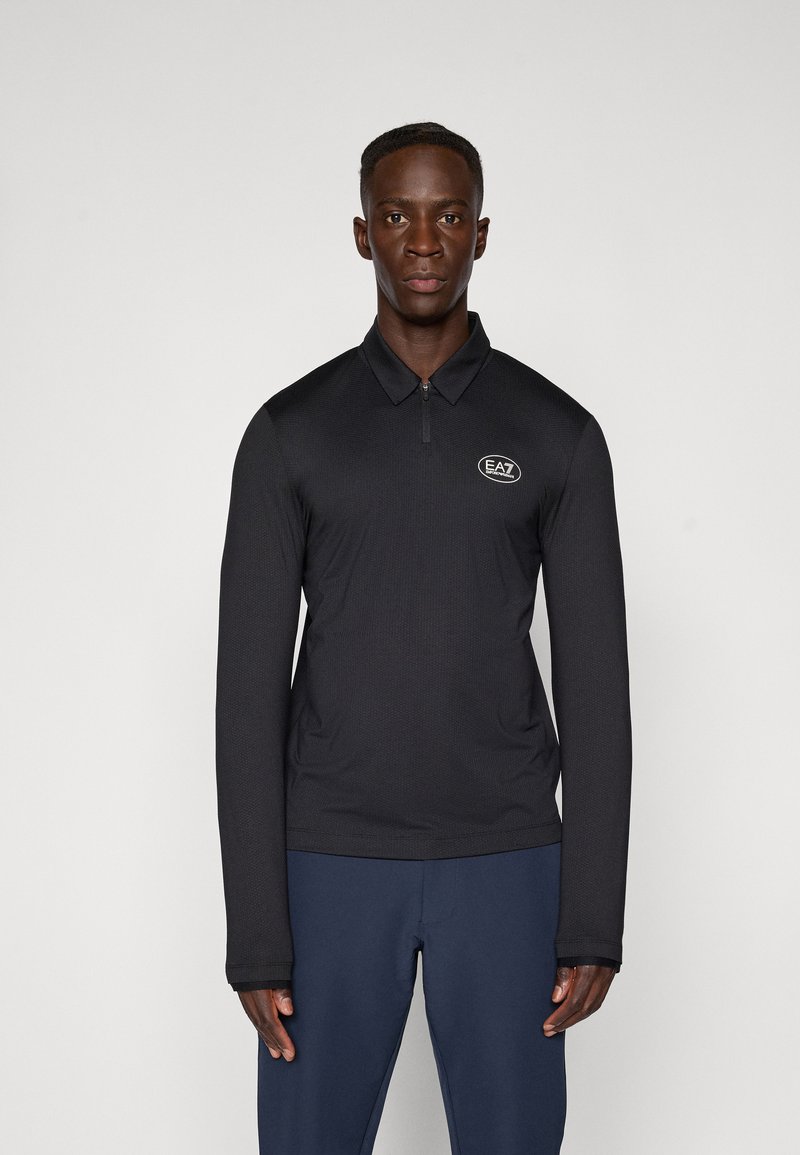 Fekete, hosszúujjú pólóing, cipzáras nyakkal, texturált anyagból készült, az EA7 logóval a mellkasán. Tengerészkék nadrággal viselve.
